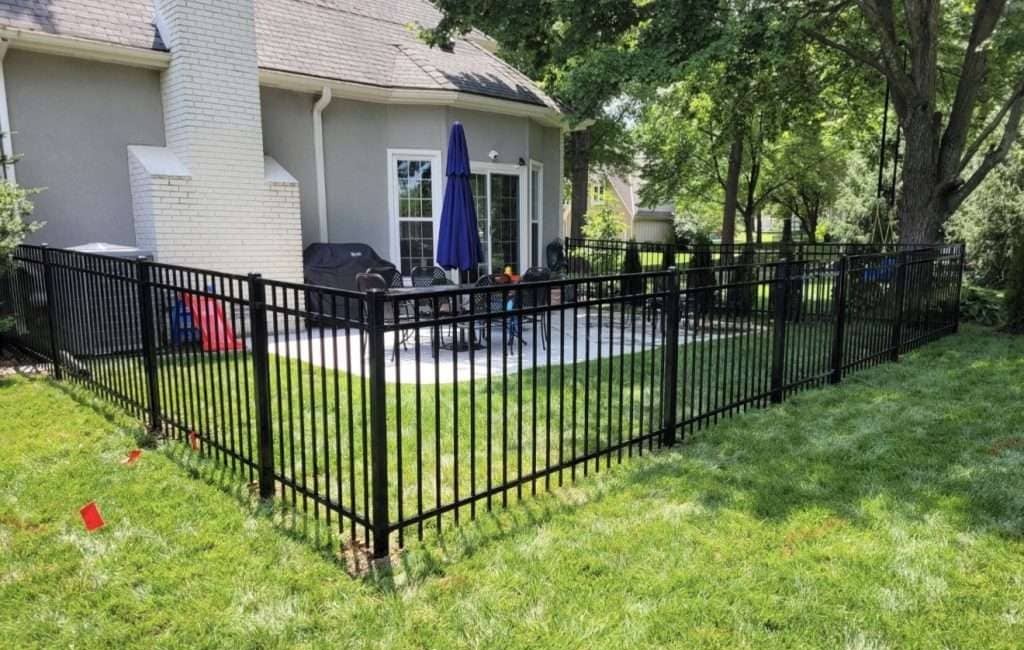 Black ornamental aluminium fence enclosing a patio and lawn behind a grey brick house.