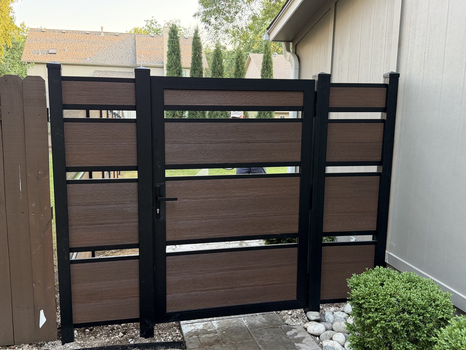 Brown horizontal-slat walk gate with black framing and narrow gaps beside a house.