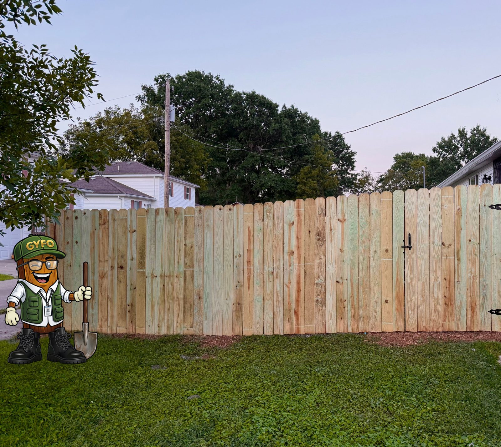 New pressure-treated wood privacy fence with a gate, site cleaned up, the GYFO mascot standing beside it with a shovel - a Get Your Fence On install