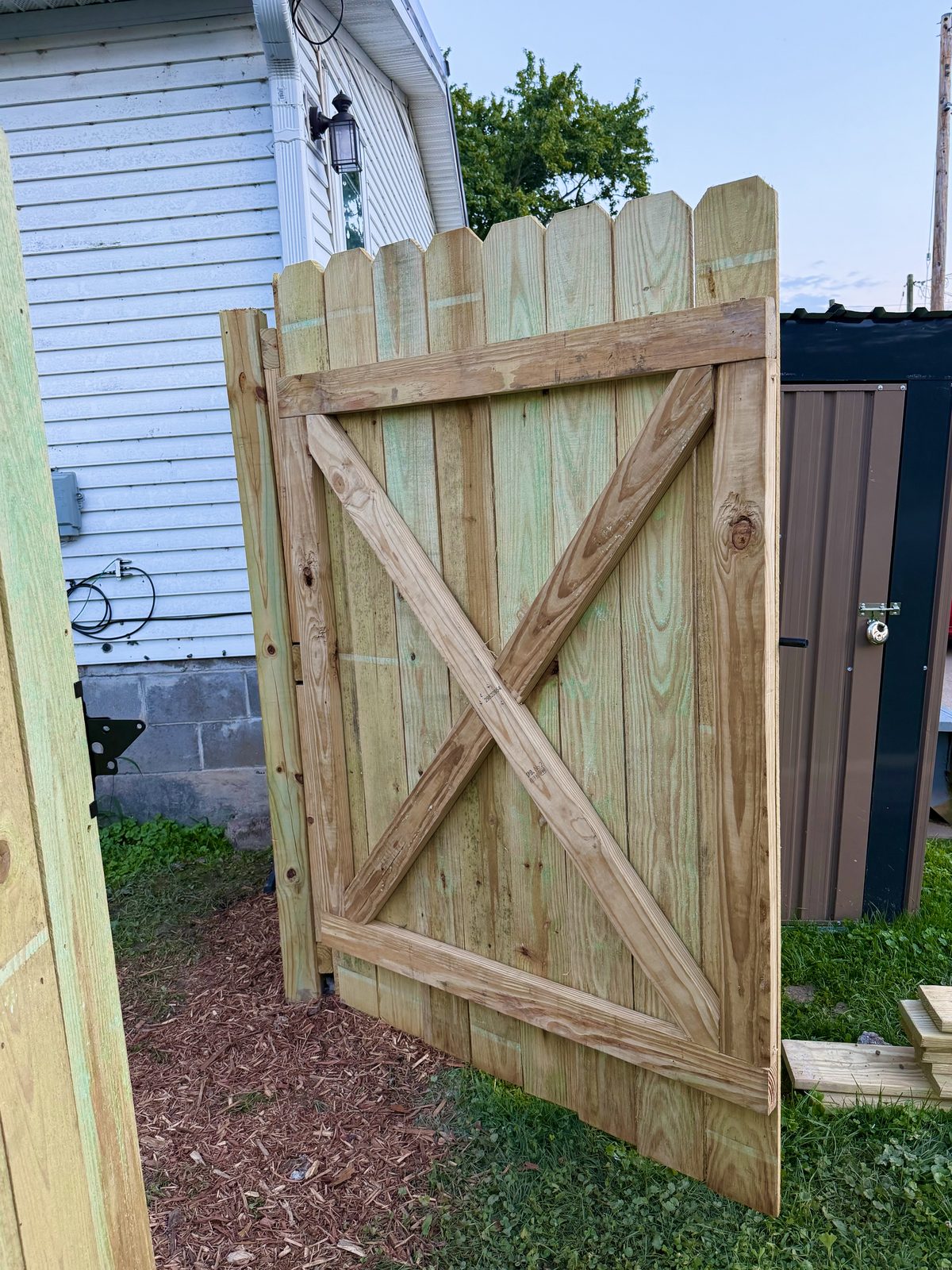 Open wood gate with dog-ear pickets and diagonal braces beside a house.