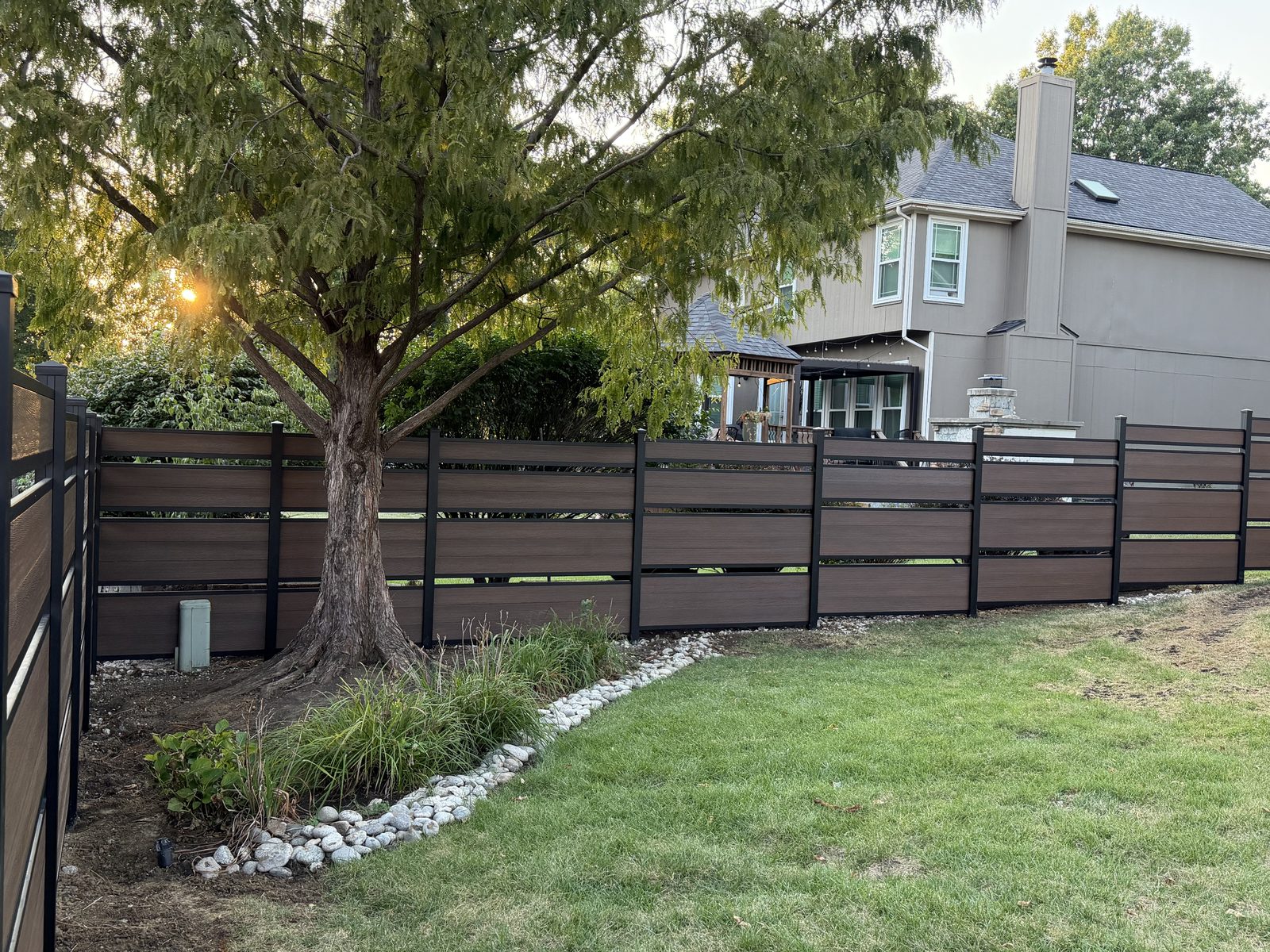 A finished brown horizontal composite fence on black posts running past a mature tree and river-rock border at golden hour
