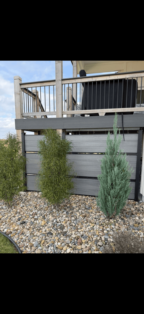 Gray horizontal screen beneath a raised deck, with shrubs in a stone bed in front.