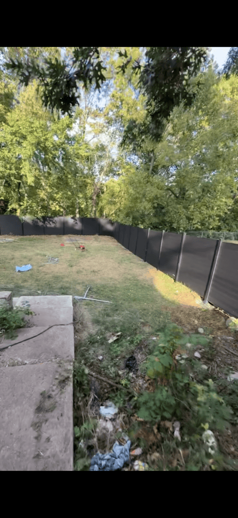 Dark privacy fence wrapping around a grassy yard beneath trees.