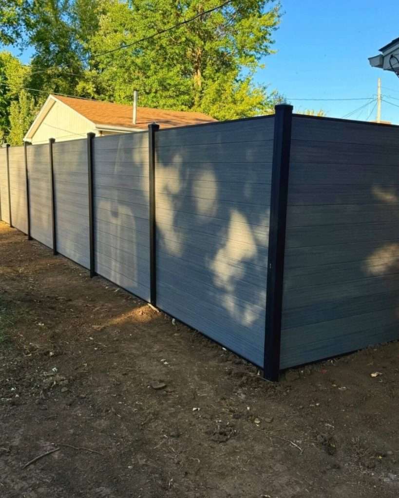 Gray horizontal privacy fence turning a corner on bare ground, with tree shadows across the panels.