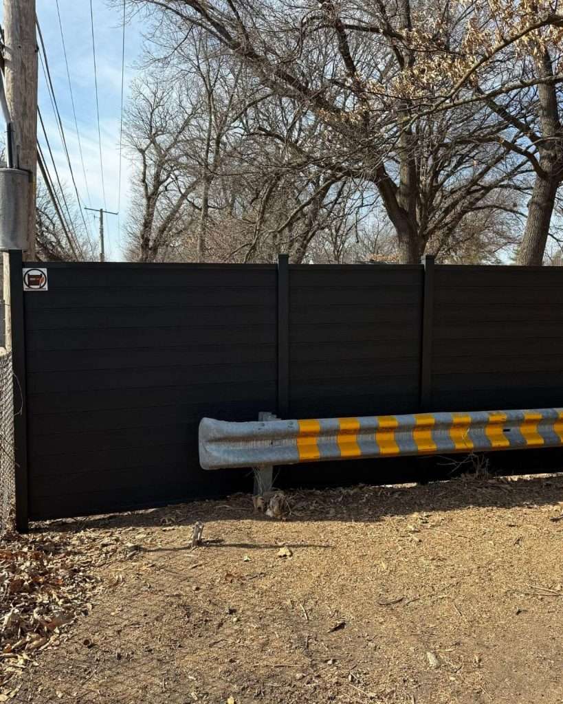 Dark horizontal privacy fence behind a roadside guardrail with yellow reflectors.
