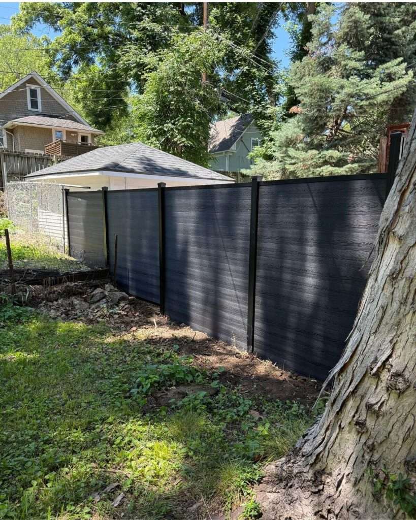 Dark horizontal privacy fence running between mature trees and a small outbuilding.