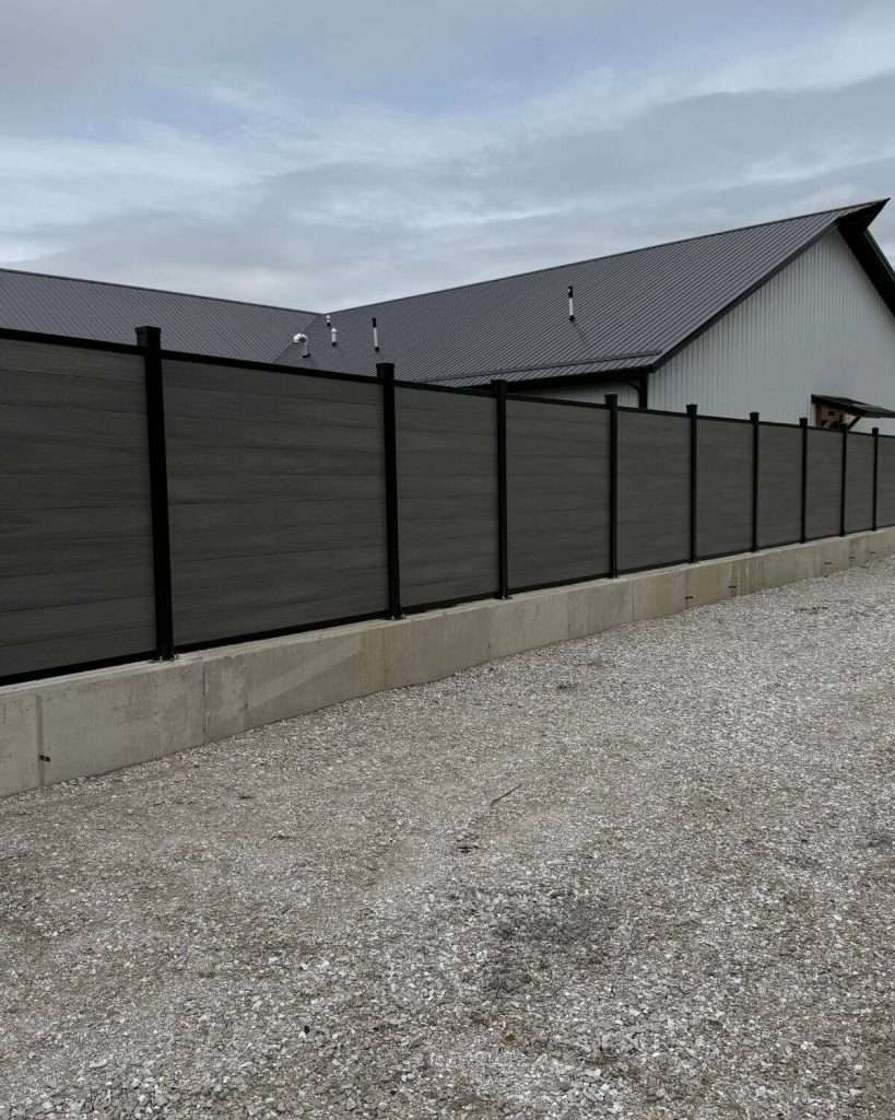 Dark horizontal privacy fence on a concrete curb beside a gravel area and outbuilding.