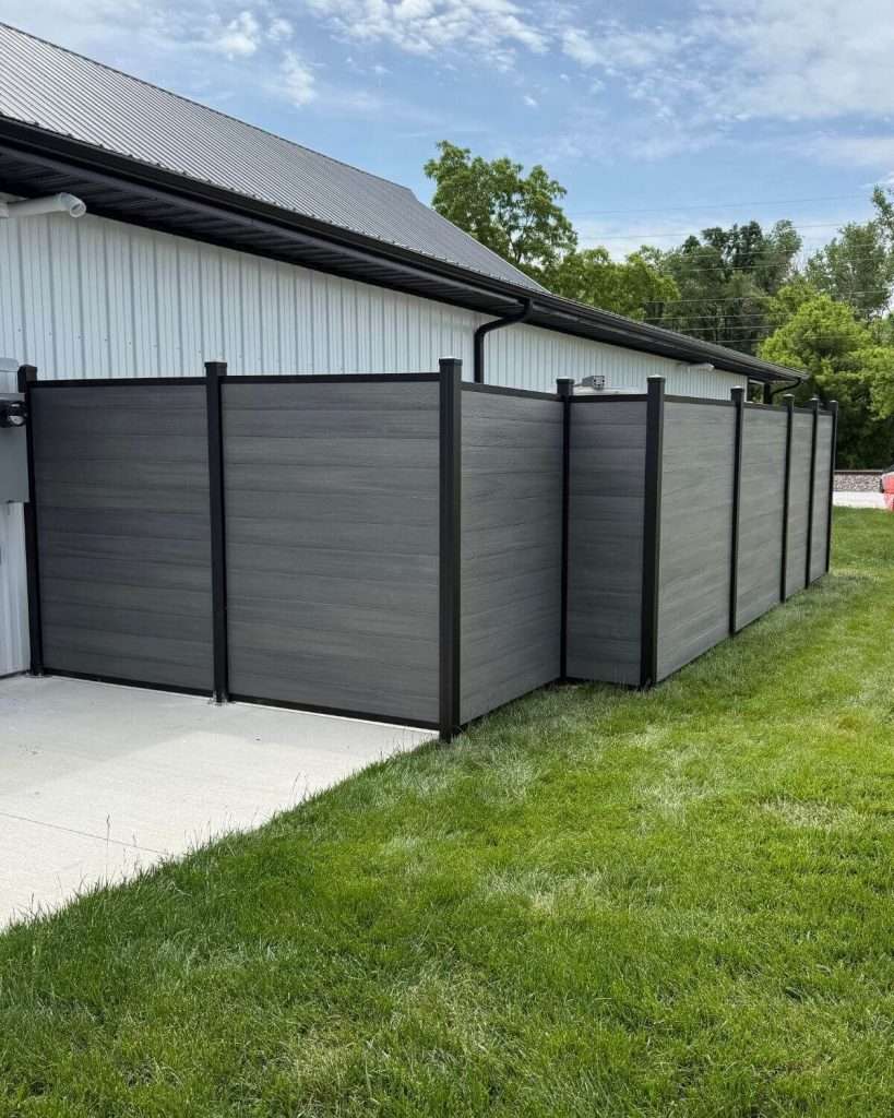 Gray horizontal fence enclosing an area beside a metal-sided building and concrete pad.