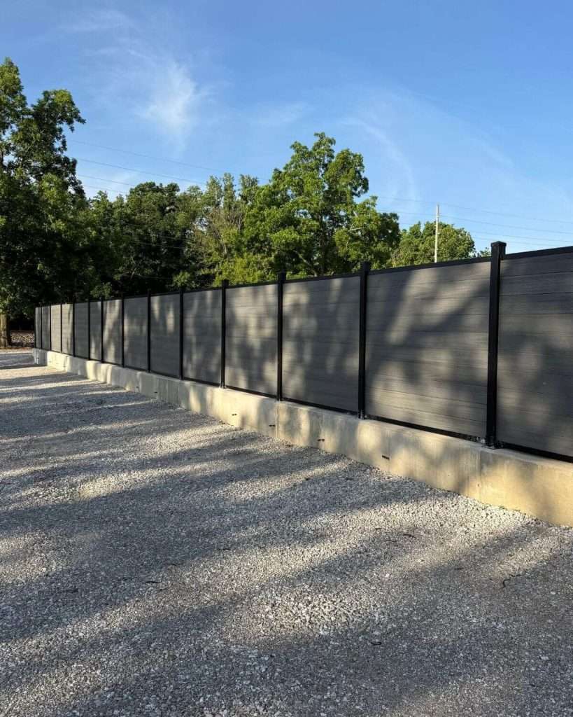 Gray horizontal privacy fence on a concrete curb beside a gravel driveway.