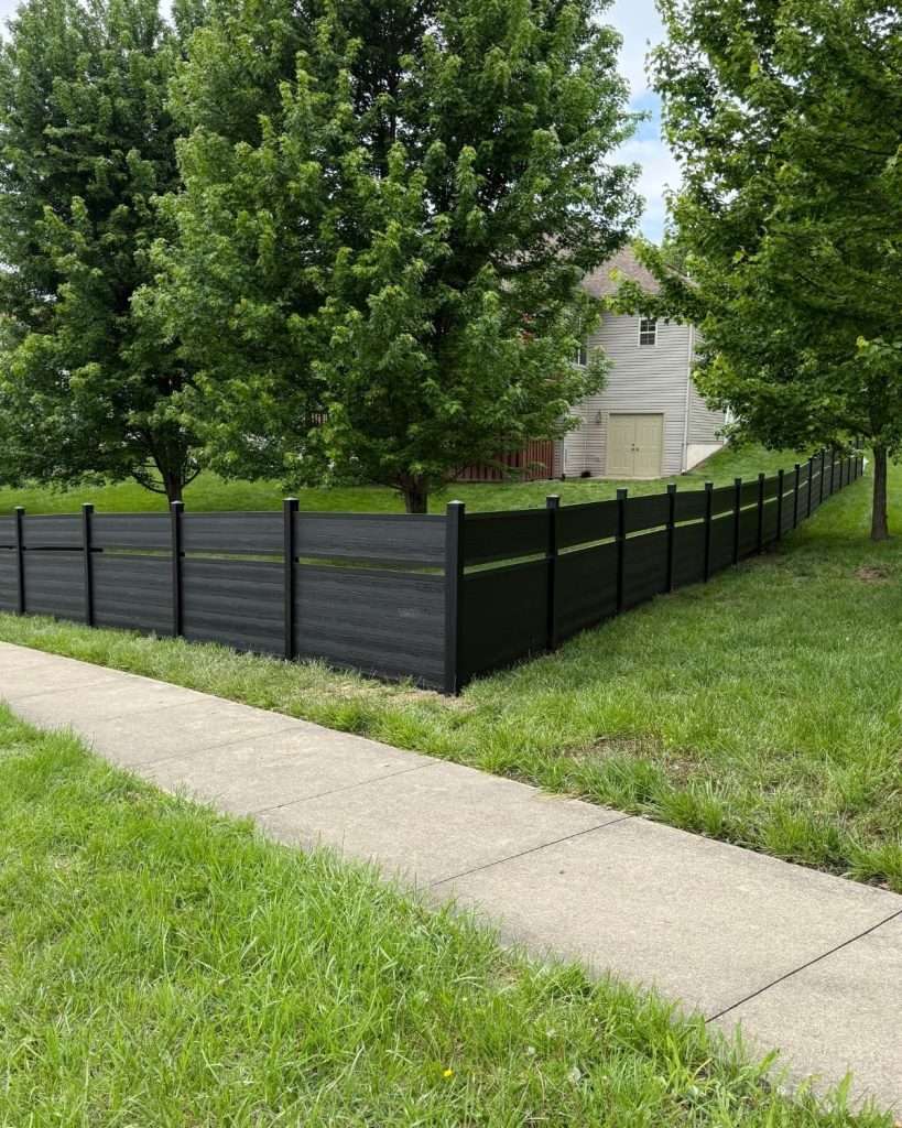 Dark horizontal fence turning a corner beside a sidewalk and tree-lined lawn.