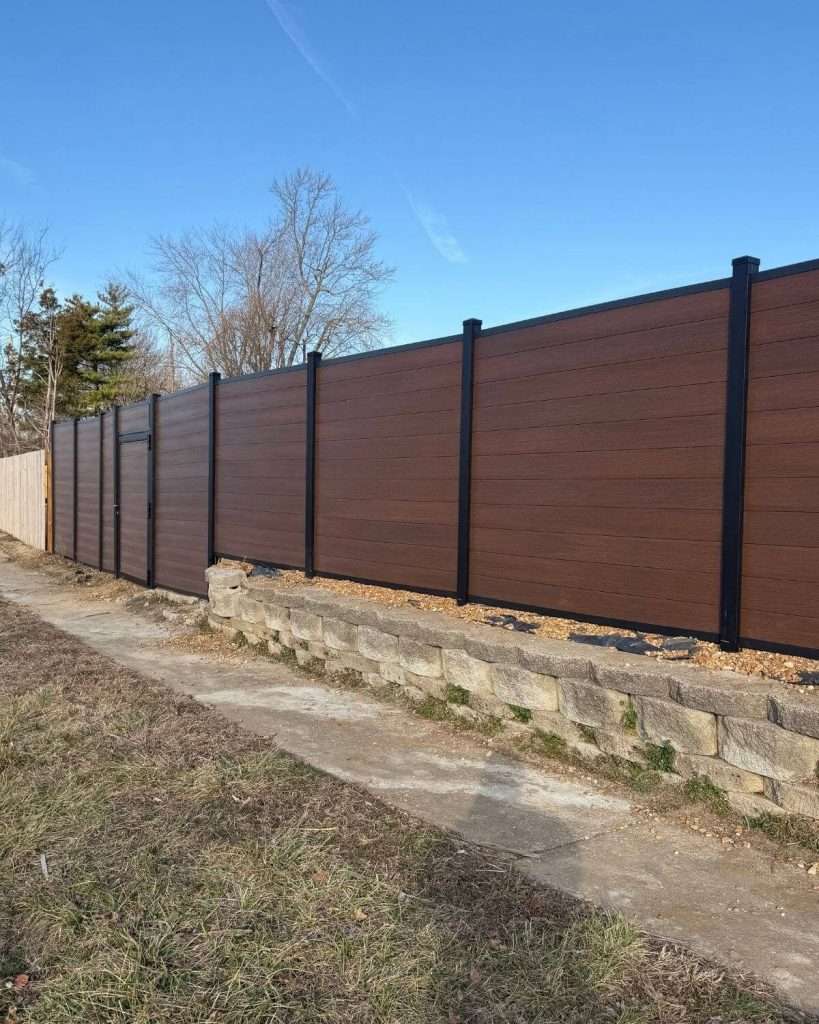 Brown horizontal privacy panels and black posts above a stone retaining wall beside a walkway.