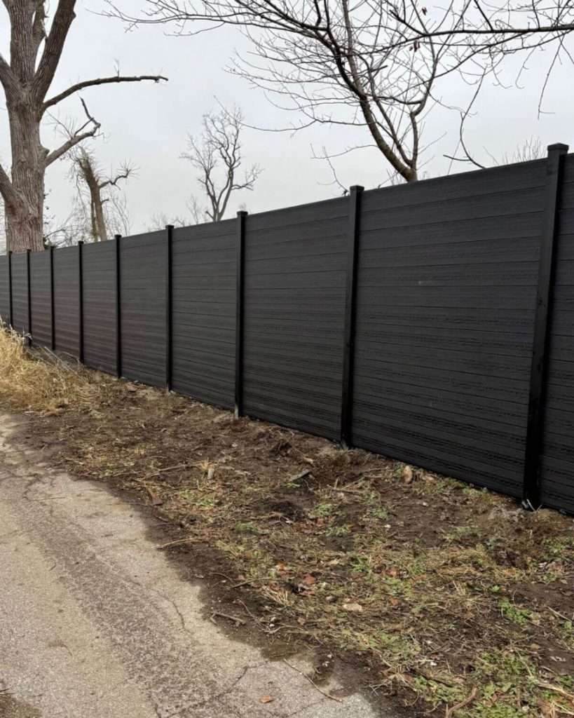 Dark horizontal privacy fence following a roadside slope beneath leafless trees.
