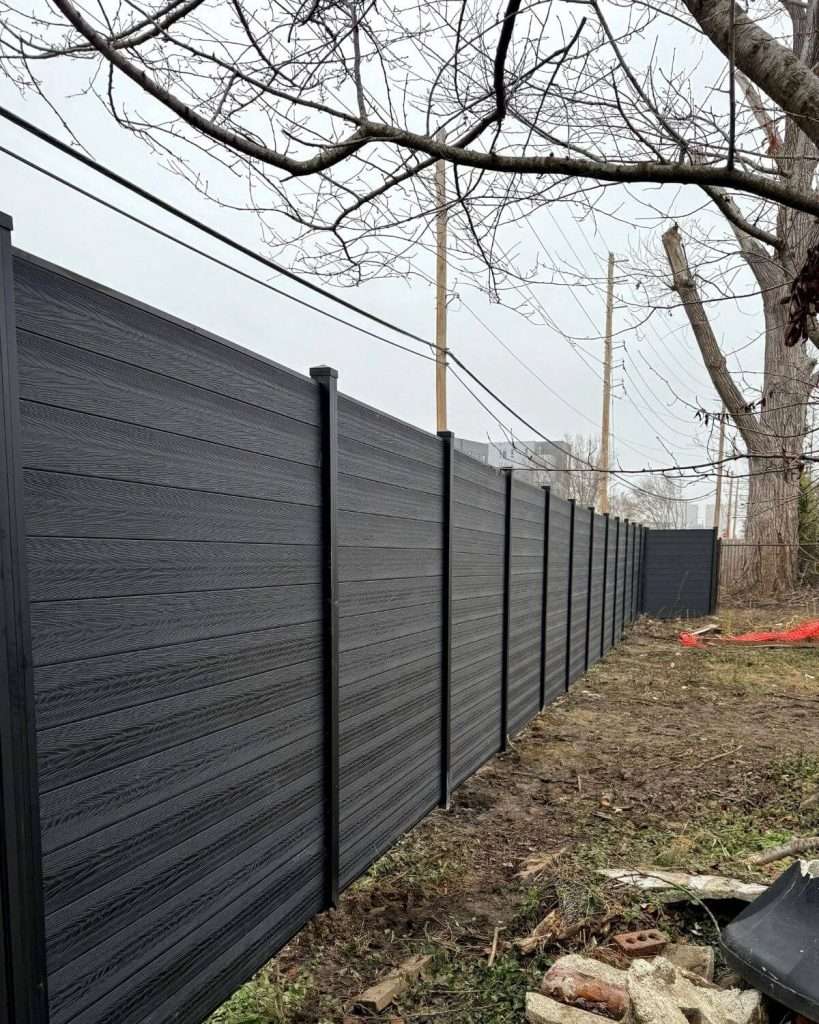 Long gray horizontal privacy fence with black posts beside trees and uneven ground.