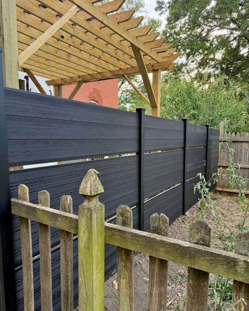 Dark horizontal privacy screen beside a timber pergola and an older wood picket fence.